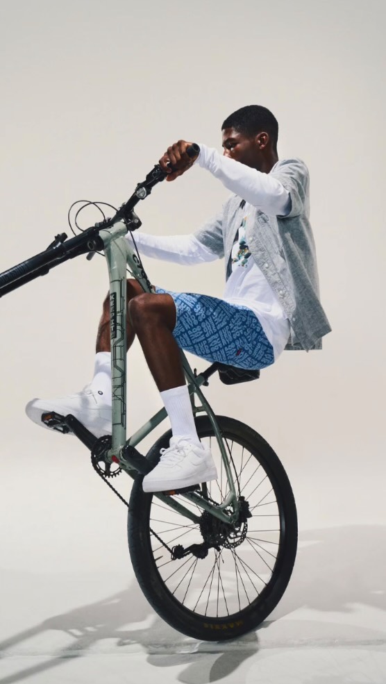 A man performs a wheelie on a mountain bike indoors. He wears casual clothing with white sneakers, showcasing balance and bike control.