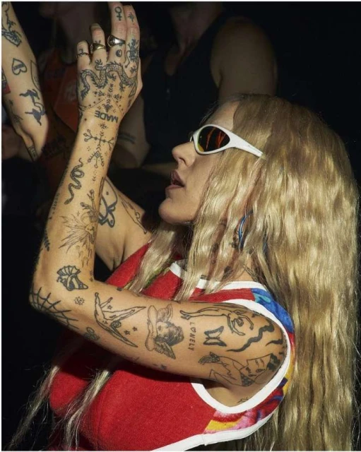 Woman with blond hair and sunglasses raises her tattooed arms indoors. She wears a red top with blue and white trim, and is at eye-level.