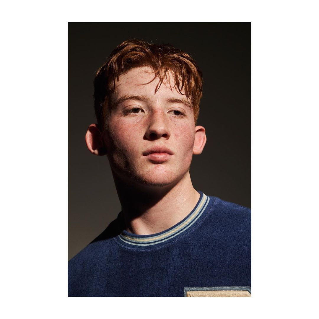 Portrait of a young man with red hair wearing a navy blue t-shirt in a photo studio with dramatic lighting and a neutral gray background.