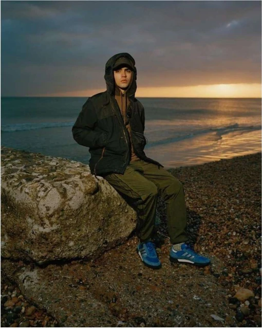 Young man in a hooded jacket sits on a rock at the beach against a cloudy sky, showcasing casual outdoor fashion and coastal scenery.