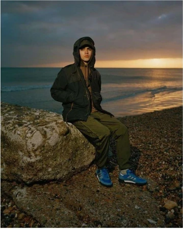 Young man in a hooded jacket sits on a rock at the beach against a cloudy sky, showcasing casual outdoor fashion and coastal scenery.