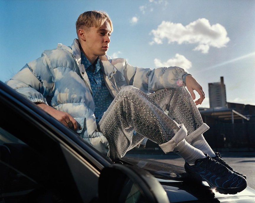 Young man in a cloud-print puffer jacket sitting on the hood of a car against a cloudy sky background.