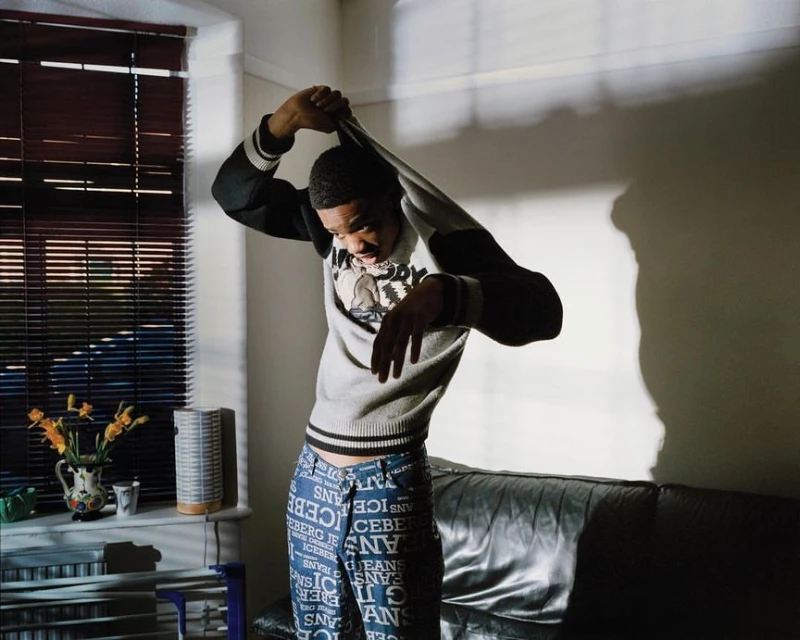 Young man removing grey sweater inside sunlit room with window blinds, flowers, and leather couch visible in the background.