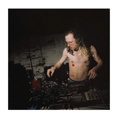 Shirtless man with long curly hair operating audio mixing equipment in a dimly lit room with direct flash.