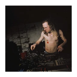 Shirtless man with long curly hair operating audio mixing equipment in a dimly lit room with direct flash.