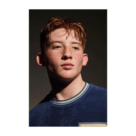 Portrait of a young man with red hair wearing a blue crewneck sweater against a dark background.