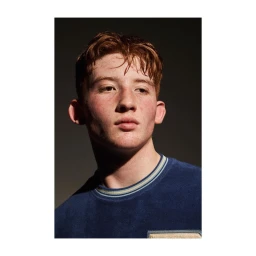 Portrait of a young man with red hair wearing a blue crewneck sweater against a dark background.