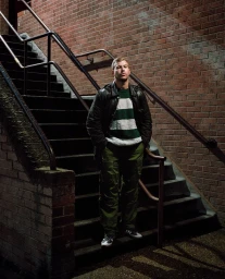 Man standing on outdoor brick staircase wearing a black leather jacket, green striped sweater, and cargo pants.