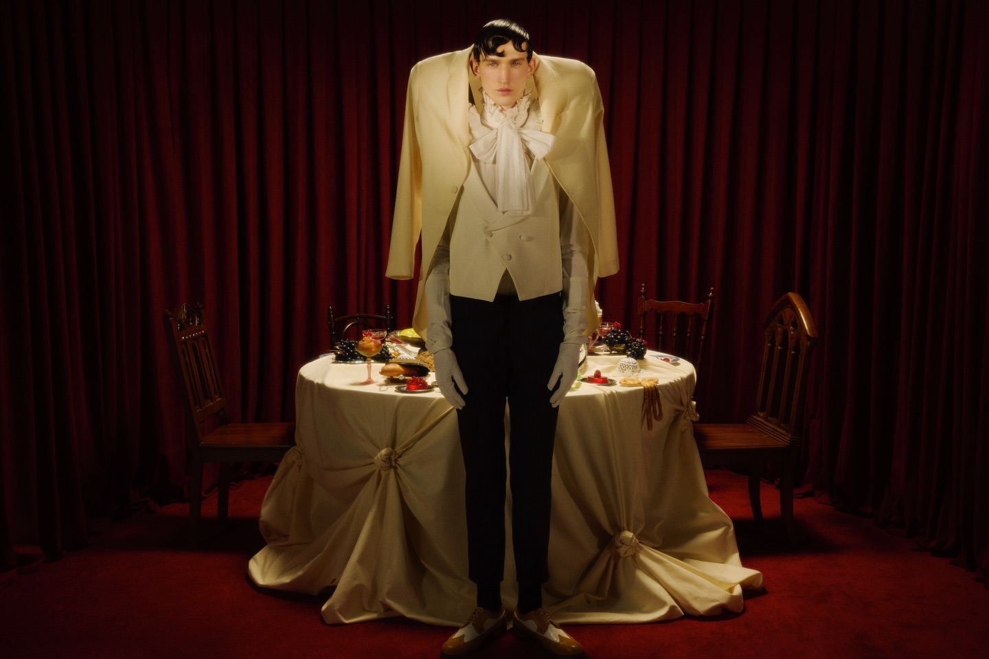 Adult male in a light beige suit jacket and black pants stands at a table with a white tablecloth, against a backdrop of red curtains.