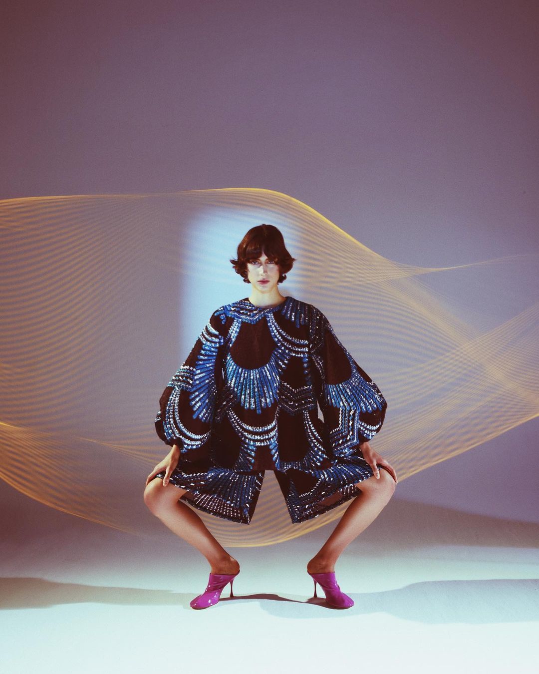 A woman poses in a fashion shot, wearing a patterned outfit and pink heels, with a dynamic light pattern in the background.