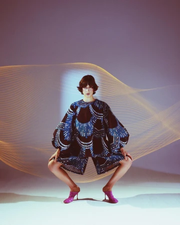 A woman poses in a fashion shot, wearing a patterned outfit and pink heels, with a dynamic light pattern in the background.