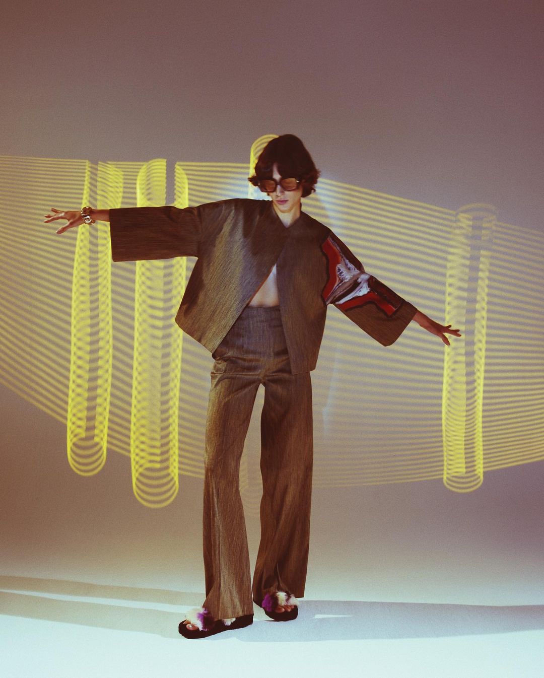 An adult female model poses in a brown two-piece outfit in a studio setting with yellow light lines in the background, captured in a full shot.