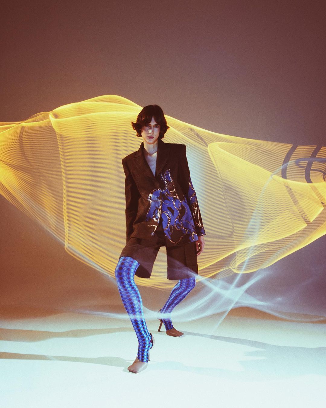 A person wearing a jacket and patterned leggings poses in front of a light painting effect backdrop with warm and cool color scheme.