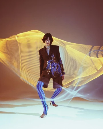 A person wearing a jacket and patterned leggings poses in front of a light painting effect backdrop with warm and cool color scheme.