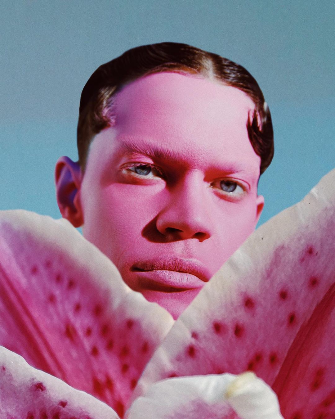 Close-up portrait of a pink-skinned adult male partially obscured by a pink lily flower against a light blue background in studio lighting.