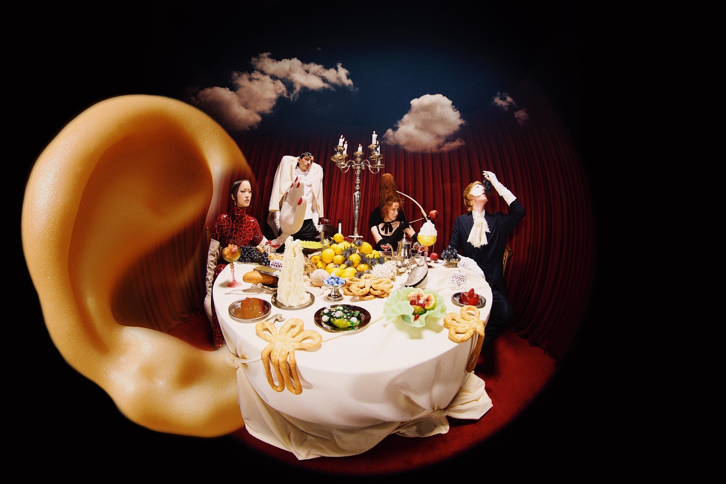 Four figures pose around a table overflowing with food, set against a backdrop of clouds and curtains in a surreal composition.