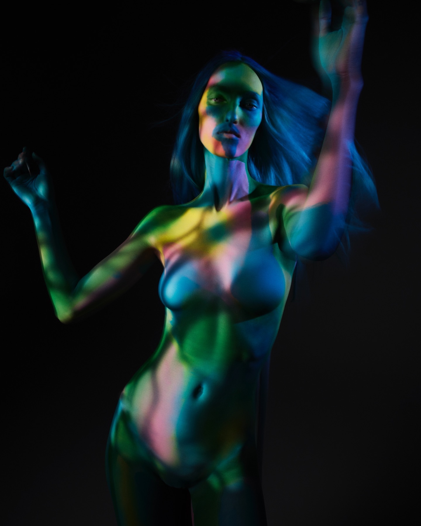 Woman with multi-colored lighting on her body stands against a dark background in a beauty style shot, showing artful color and creative lighting.