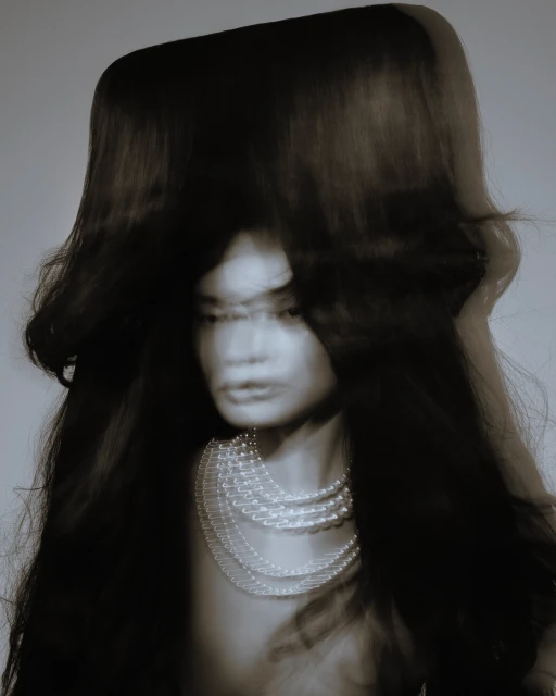 Blurred portrait of a woman with long black hair wearing a pearl necklace against a plain background in a fashion style image.