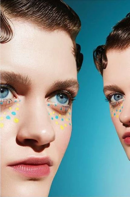 Close-up beauty shot of two fair-skinned adult females with blue eyes, styled hair, light pink lipstick and yellow/blue dots on their faces.