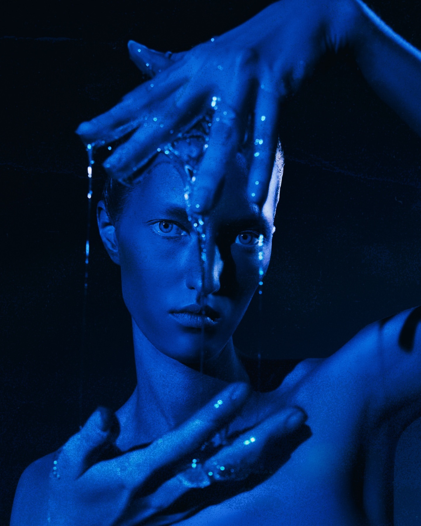 Adult female with blue glitter poses in a photo studio, under blue lighting, with hands framing face, dark background.