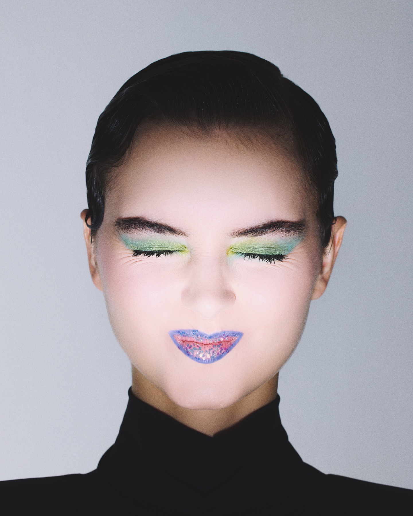 Close-up portrait of a woman with closed eyes, wearing vivid green eyeshadow and vibrant blue lipstick with reddish accents on her lips.