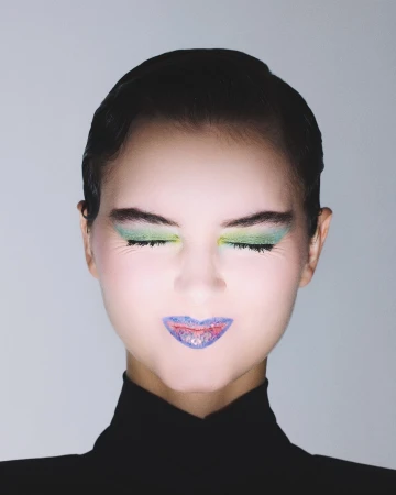 Close-up portrait of a woman with closed eyes, wearing vivid green eyeshadow and vibrant blue lipstick with reddish accents on her lips.