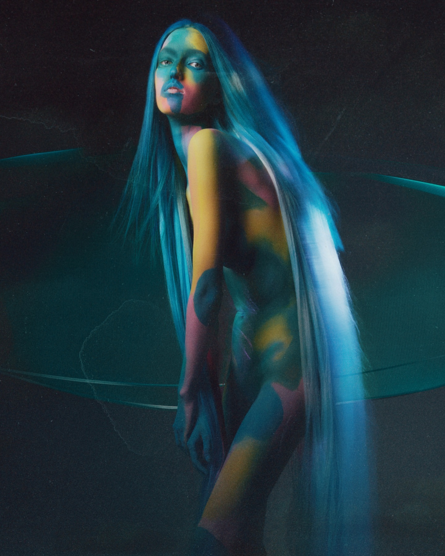 An androgynous fashion model with blue hair and colorful body paint poses in a medium studio shot with dark tones and soft lighting.