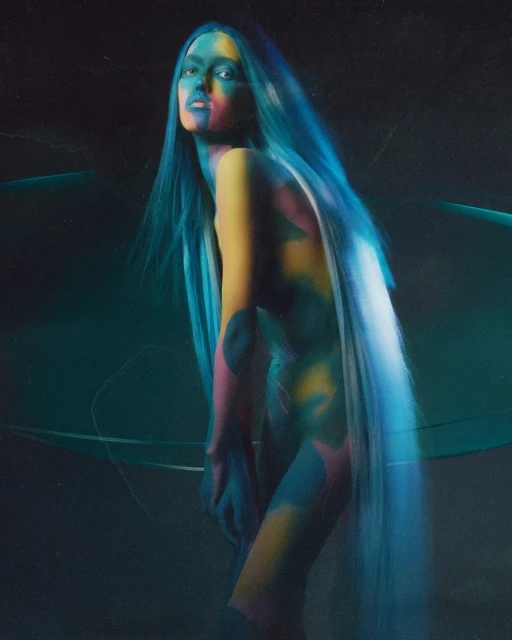 An androgynous fashion model with blue hair and colorful body paint poses in a medium studio shot with dark tones and soft lighting.