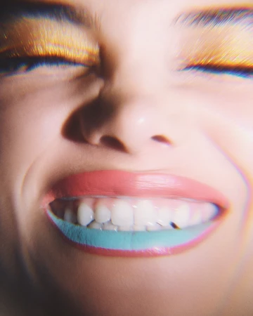Close-up shot of a woman smiling, showcasing her teeth and brightly colored makeup, including golden eye shadow and two-toned lip color.