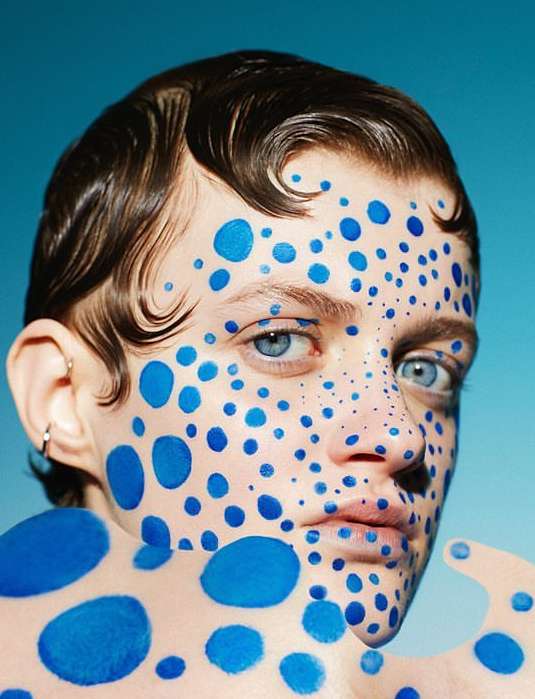 Close-up of a young adult with blue eyes and short brown hair, covered in blue dots, against a solid blue background.
