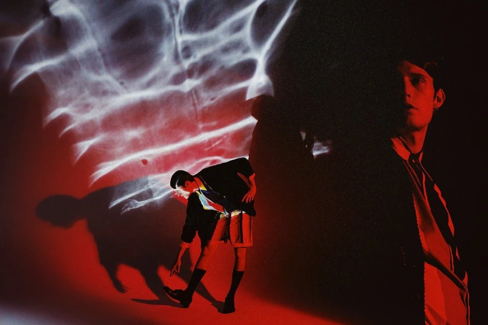 Person in shadow, contrasted with water reflection. A man poses indoors in shorts and a jacket, one arm reaching toward the ground.