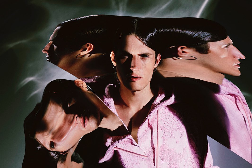 Portrait of a man with slicked-back hair wearing a pink robe, repeated and distorted on a patterned, grey background, indoor shot.