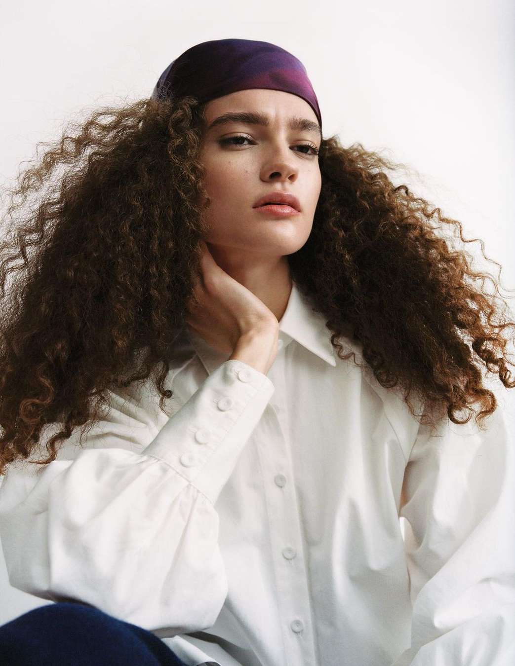 Portrait of a young adult woman with curly hair wearing a purple headscarf and white collared shirt against a white background.