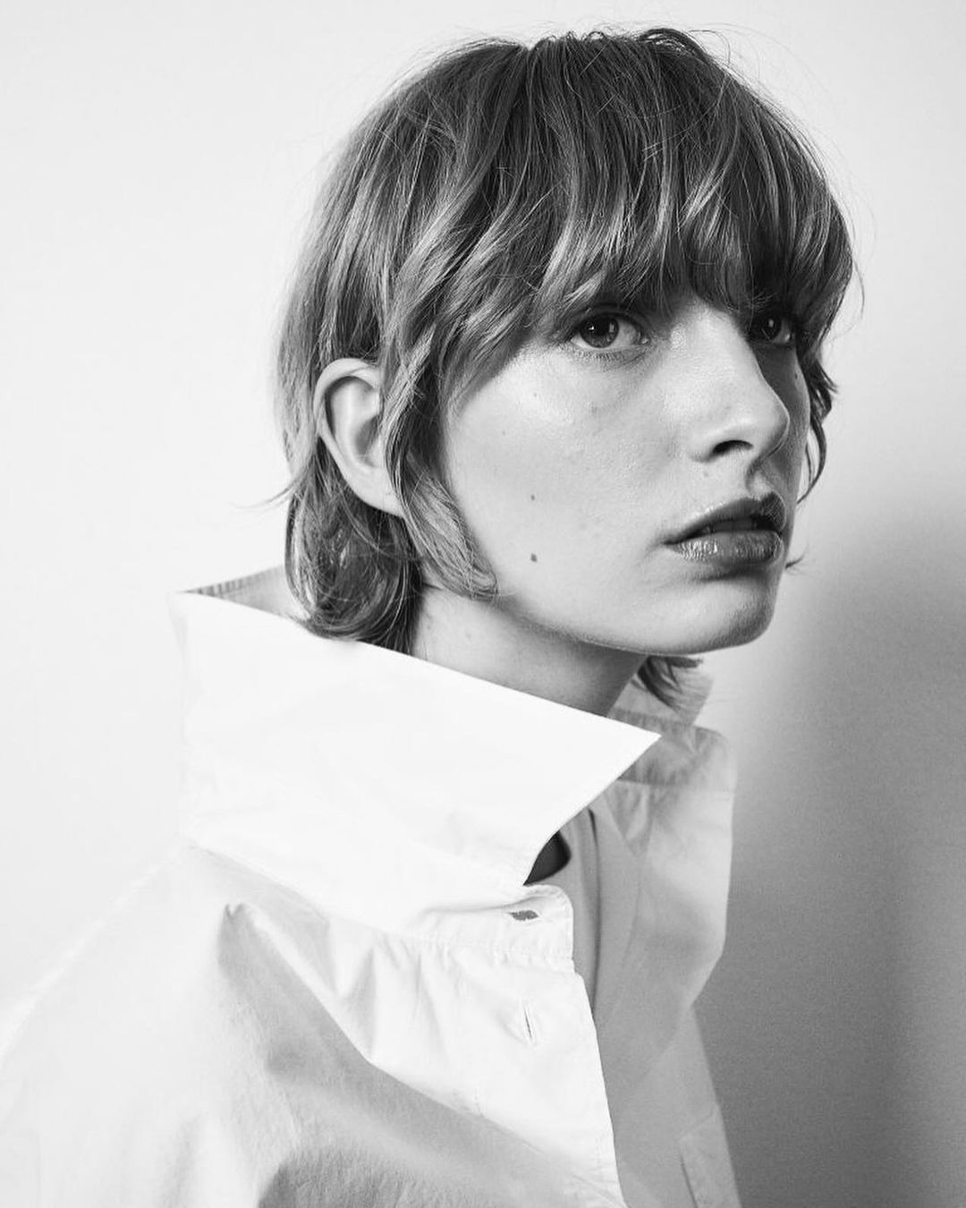 Black and white portrait of a person with short wavy hair, wearing a white collared shirt against a neutral background.