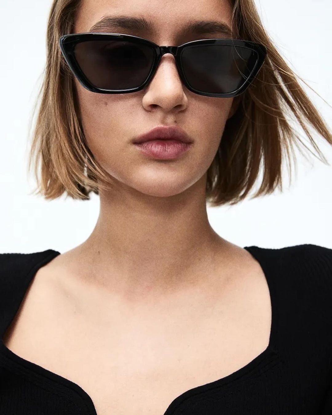 Close-up of a woman with short brown hair and black sunglasses, wearing a black top against a bright background, fashion style.