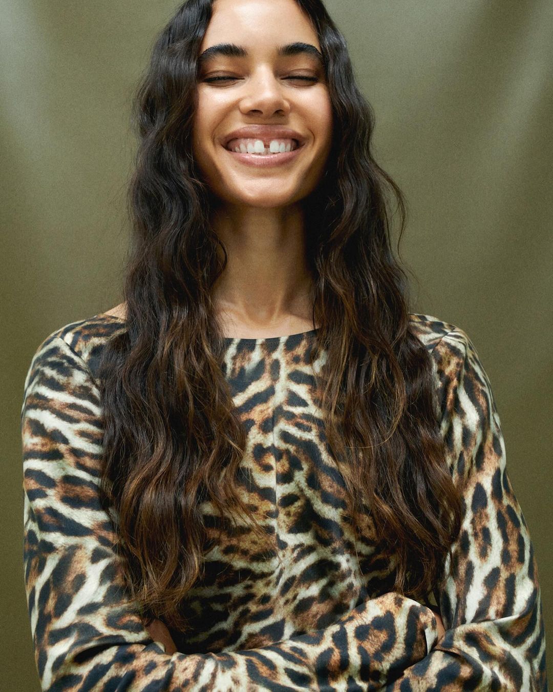 Smiling woman with long brunette hair and a leopard print top posing against a solid olive green background in a medium close-up shot.