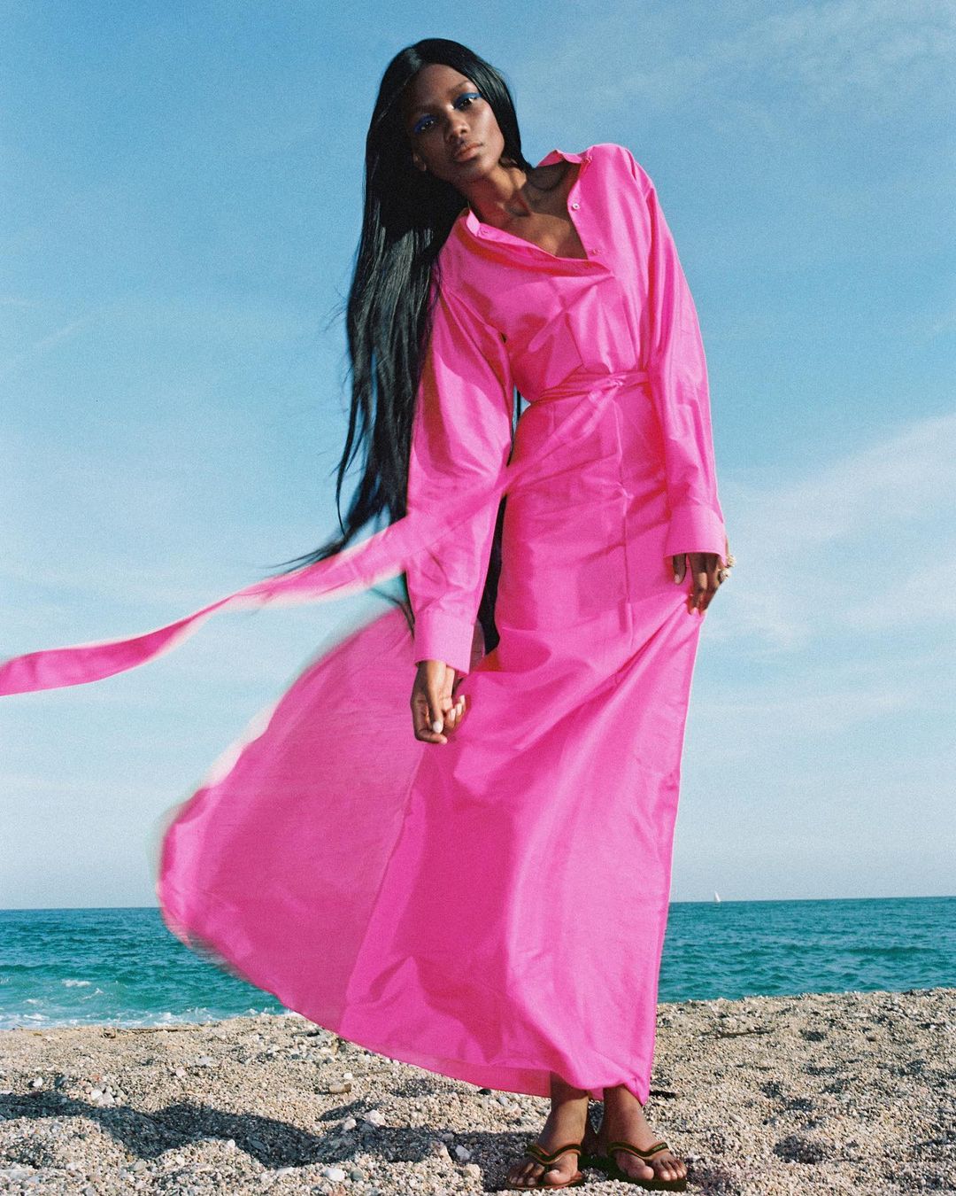 A woman in a bright pink dress stands on a sandy beach with the ocean and sky visible in the background, creating a vibrant fashion image.