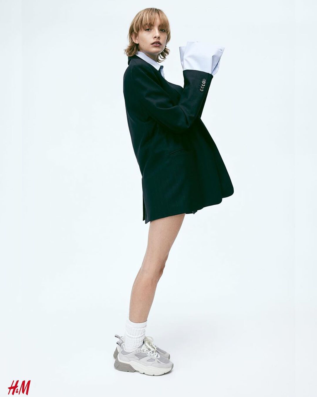 A slender female model with short blonde hair poses in a photo studio wearing a navy blazer, long-sleeve shirt and gray sneakers against a white backdrop.
