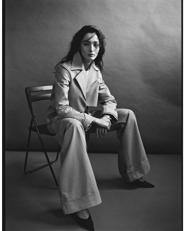 Black and white portrait of a woman seated on a folding chair wearing a tailored jacket and wide-leg trousers
