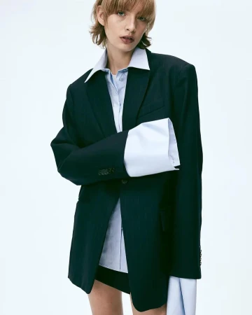 Model wearing an oversized dark blazer over an untucked light blue button-down shirt with long extended cuffs against a plain white background