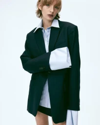 Model wearing an oversized dark blazer over an untucked light blue button-down shirt with long extended cuffs against a plain white background