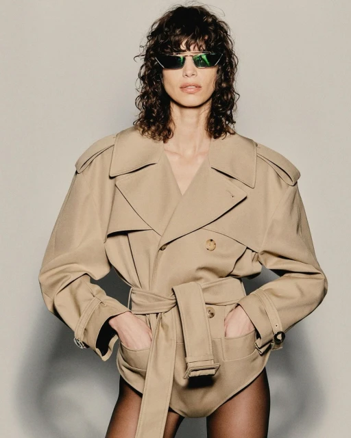A woman poses in a belted trench coat and black tights against a plain background, highlighted by her curly hair and sunglasses.