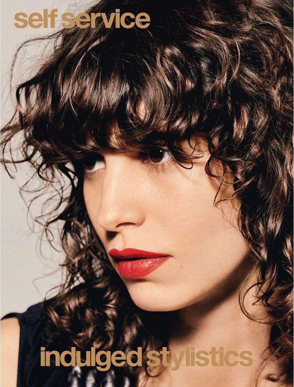 Close-up portrait of a woman with dark curly hair and bright red lipstick against a neutral grey background.