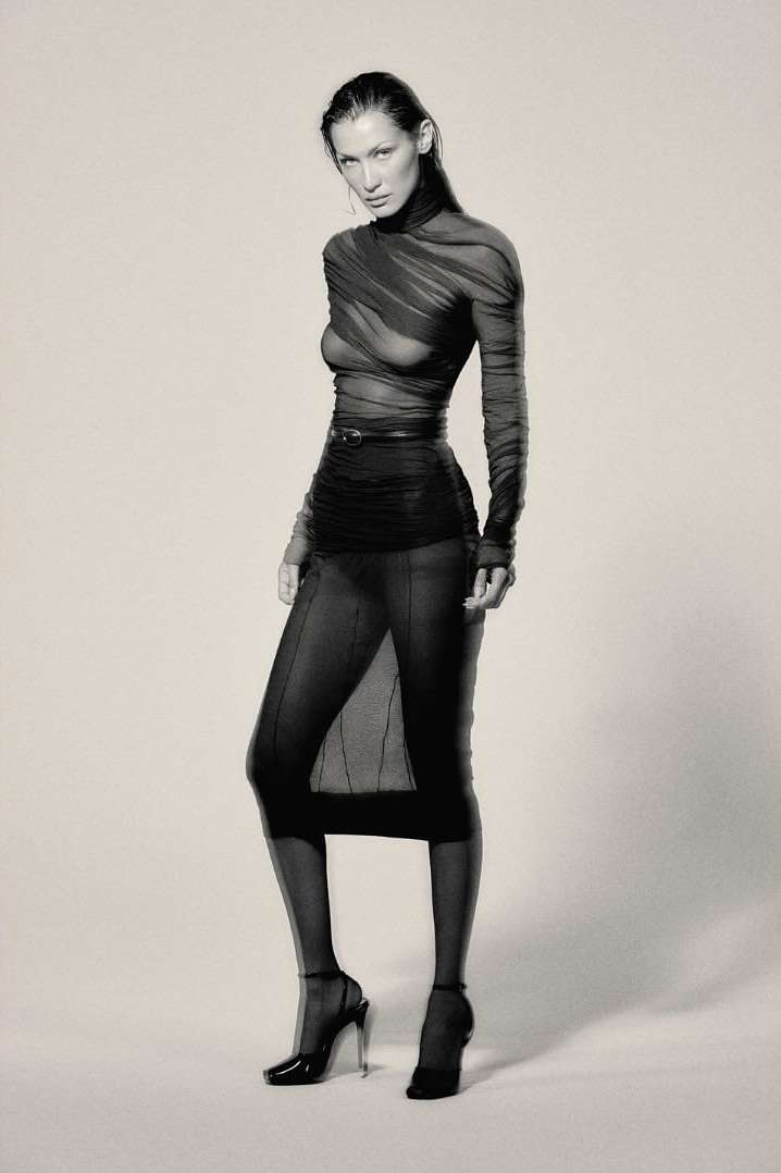 Studio shot of a young adult woman in a sheer black long-sleeve top, skirt, and high heels posing against a neutral background.