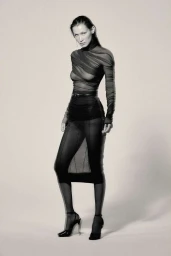 Bella Hadid in a sheer black ruched turtleneck top and skirt with black heels against a light background