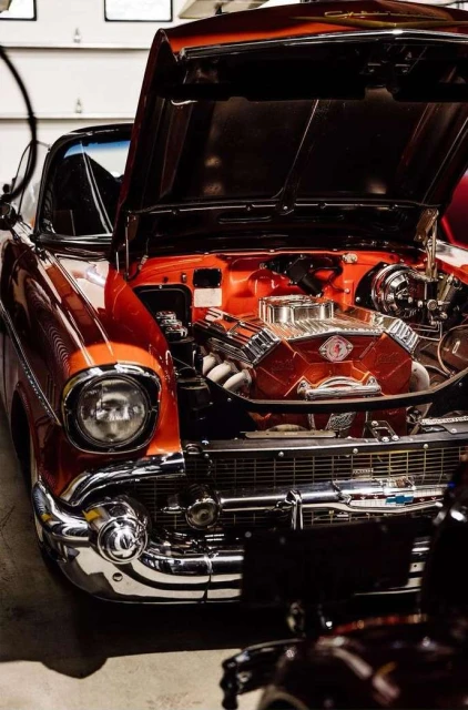 Close-up of a classic orange and chrome car with its hood open, showcasing the engine; shot indoors with soft, diffused lighting.