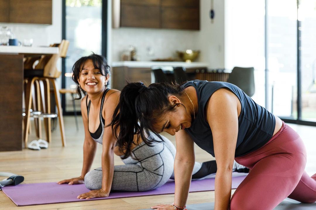 Two adult women stretch on yoga mats in a bright interior. One woman smiles at the camera; the other leans forward in a stretch.