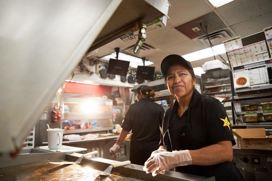 A woman in a work uniform stands in a commercial kitchen, with another woman working in the background, and an overhead shot.