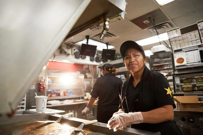 A woman in a work uniform stands in a commercial kitchen, with another woman working in the background, and an overhead shot.