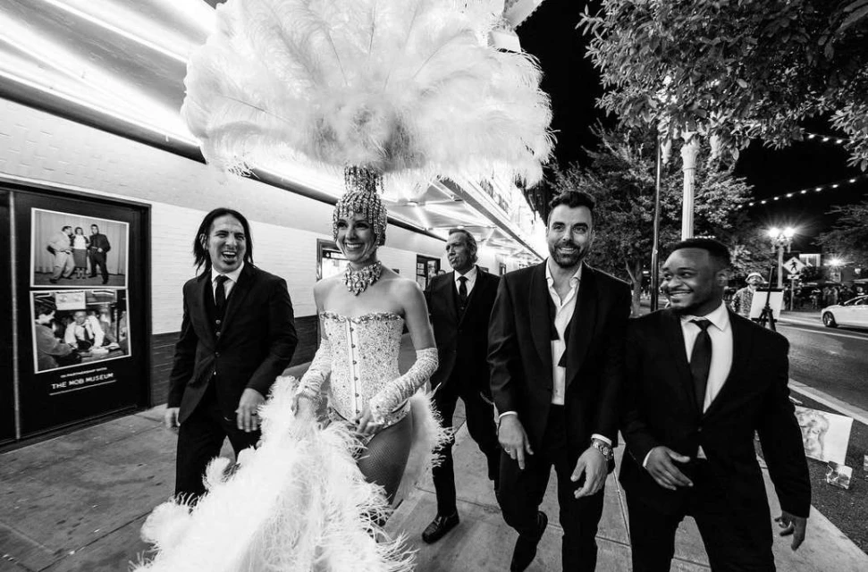 Black and white outdoor shot of five adults: a woman in a feather headpiece and men's suits, standing in front of a building at night.